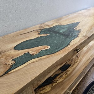 K&ouml;nnte beinhalten: Ein langer Holzkonsolentisch mit einer hellen Holzmaserung und einer gro&szlig;en, unregelm&auml;&szlig;igen Einlage aus durchscheinendem, blaugr&uuml;nem Epoxidharz. Der Tisch verf&uuml;gt &uuml;ber eine untere Ablage mit &auml;hnlichen Holz- und dunklen Harzdetails.