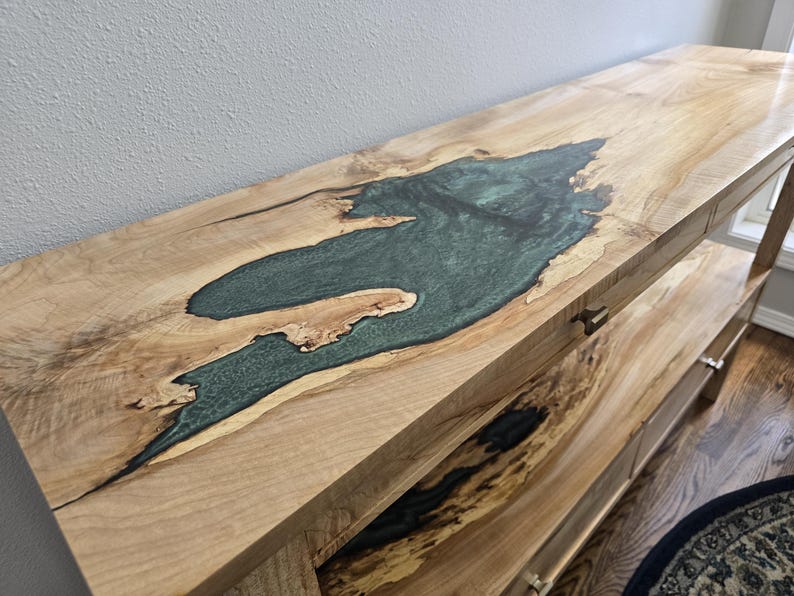 K&ouml;nnte beinhalten: Ein langer Holzkonsolentisch mit einer hellen Holzmaserung und einer gro&szlig;en, unregelm&auml;&szlig;igen Einlage aus durchscheinendem, blaugr&uuml;nem Epoxidharz. Der Tisch verf&uuml;gt &uuml;ber eine untere Ablage mit &auml;hnlichen Holz- und dunklen Harzdetails.