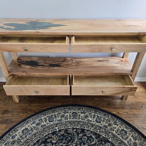 K&ouml;nnte beinhalten: Ein Konsolentisch aus hellem Holz mit nat&uuml;rlicher Baumkante, vier offenen Schubladen mit kleinen quadratischen Metallgriffen und einer unteren Ablage mit dunkler, unregelm&auml;&szlig;iger Harzeinlage. Die Tischplatte zeigt ebenfalls eine dunkle Harzeinlage.