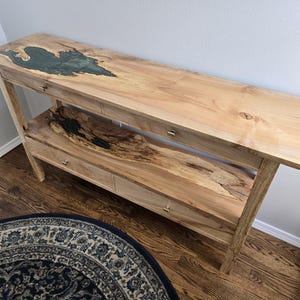 K&ouml;nnte beinhalten: Ein langer Holzkonsolentisch mit einer nat&uuml;rlichen hellen Holzmaserung. Die Oberfl&auml;che enth&auml;lt einen eingelegten dunkelgr&uuml;nen Harzabschnitt, w&auml;hrend das untere Regal eine markante, dunkle und strukturierte Holzmaserung mit einer tiefschwarzen Harzeinlage aufweist.