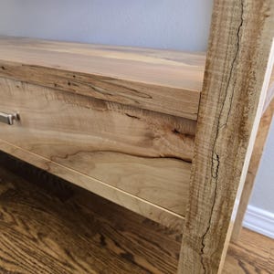K&ouml;nnte beinhalten: Ein Konsolentisch aus hellem Holz mit einer einzelnen Schublade und einem kleinen, rechteckigen Griff aus silberfarbenem Metall. Das Holz zeigt eine nat&uuml;rliche Maserung mit dunklen Streifen und einem Riss im seitlichen Rahmen.