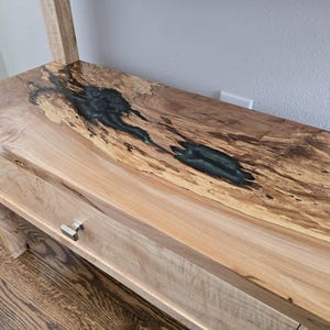 K&ouml;nnte beinhalten: Ein Holzregal mit einer nat&uuml;rlichen Baumkantenoberfl&auml;che und einer dunkelgr&uuml;nen Harzeinlage, die die Holzmaserung ausf&uuml;llt. Das Regal verf&uuml;gt &uuml;ber eine glatte, helle Holzschubladenfront, die mit einem kleinen Metallgriff ausgestattet ist.