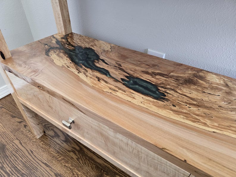 K&ouml;nnte beinhalten: Ein Holzregal mit einer nat&uuml;rlichen Baumkantenoberfl&auml;che und einer dunkelgr&uuml;nen Harzeinlage, die die Holzmaserung ausf&uuml;llt. Das Regal verf&uuml;gt &uuml;ber eine glatte, helle Holzschubladenfront, die mit einem kleinen Metallgriff ausgestattet ist.