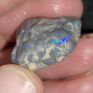 Può includere: Una pietra di opale grezza, grigia, con lampi iridescenti blu e verdi. La pietra ha una superficie strutturata ed è tenuta tra le dita, mostrando il suo stato naturale e non lucidato. Una gemma naturale unica.