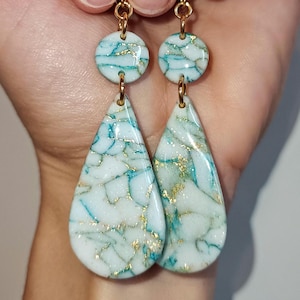 May include: A pair of dangle earrings with a teardrop-shaped design. The earrings are made of white resin with gold and teal accents. The teardrop-shaped resin is marbled with gold and teal.