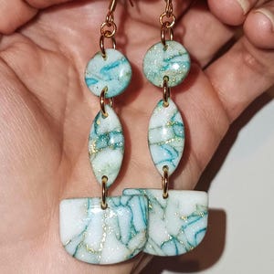 Puede incluir: Pendientes de aro dorados con cuentas de resina marmoleada en blanco y azul. Las cuentas tienen un acabado con purpurina dorada.