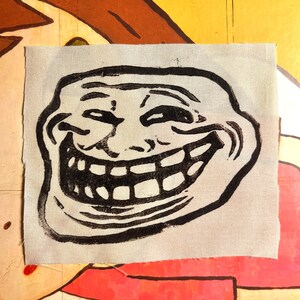 May include: A black and white graphic of the Trollface meme printed on a light gray fabric patch. The patch is rectangular with a rough edge. The background is a colorful painted surface with yellow, brown, red, and white colors.