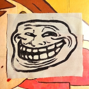 May include: A black and white Trollface meme graphic printed on a light grey fabric patch. The patch has a rectangular shape with a rough edge. The background is a colourful, painted surface.