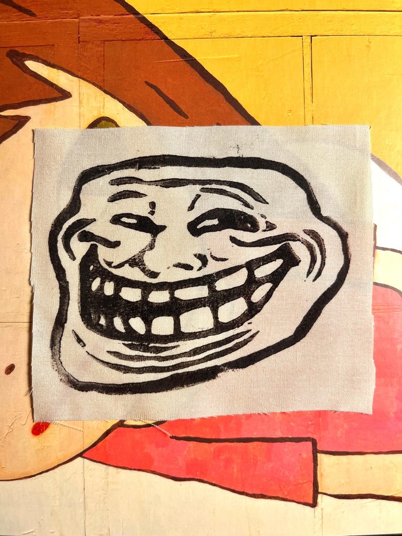 May include: A black and white graphic of the Trollface meme printed on a light gray fabric patch. The patch is rectangular with a rough, unfinished edge. The background is a colorful, painted surface.