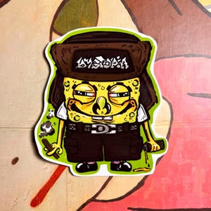 Peut inclure: Autocollant découpé représentant un personnage de dessin animé au visage jaune, portant un chapeau de fourrure marron avec le mot "AYSTOKIA" en blanc, une chemise blanche et un short marron. Le personnage tient un bâton et une bouteille.