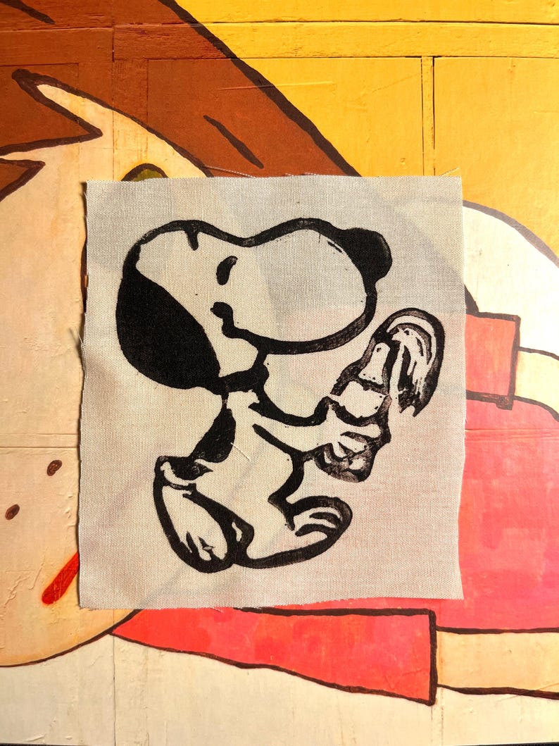 May include: A square, off-white fabric patch featuring a black illustration of Snoopy from the Peanuts comic strip. Snoopy is depicted in a seated position, holding a bottle. The background is a mix of yellow, brown, and pink.