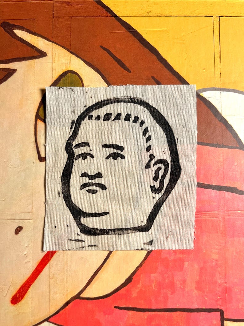 May include: A square, off-white fabric patch featuring a black linocut print of a stylized human face. The face has a rounded shape, with a prominent nose, small eyes, and a defined ear. The background is a colorful, abstract painting.