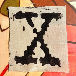 Peut inclure: Un patch de tissu carr&eacute; blanc cass&eacute; avec un grand "X" noir imprim&eacute;. Le "X" est stylis&eacute; avec des lignes &eacute;paisses et irr&eacute;guli&egrave;res, donnant un aspect imprim&eacute; &agrave; la main ou en bloc. Le fond est un collage color&eacute;.
