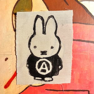 Op de afbeelding: Een kleine, rechthoekige stoffen patch met een zwart-wit cartoon konijn ontwerp. Het konijn draagt een zwarte outfit met een witte cirkel en een zwarte letter "A" erin. De patch staat op een getextureerde, gebroken witte achtergrond.