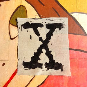 Può includere: Un piccolo pezzo quadrato di tessuto bianco sporco con una grande lettera "X" nera stampata sopra. Il tessuto è leggermente sfilacciato ai bordi. Lo sfondo presenta un'illustrazione colorata in stile cartone animato con sfumature di giallo, arancione e rosso.