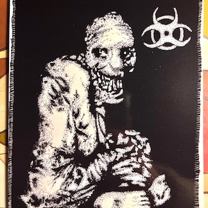 May include: Black and white patch featuring a grotesque, zombie-like figure with exposed teeth and a biohazard symbol. The image is created with a stippling technique, giving it a textured appearance. The patch has a stitched border.