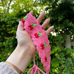 May include: A pink crochet headband with a scalloped edge, adorned with red cherries and green leaves. The headband has a tie closure. The background is a blurred view of green foliage.