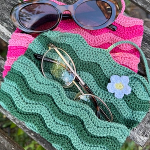 May include: Oval sunglasses with a tortoiseshell pattern sit on a pink and white striped crochet bag. Below, a green crochet bag holds gold-rimmed glasses. A small blue and yellow crochet flower decorates the green bag. The items are on a wooden surface.
