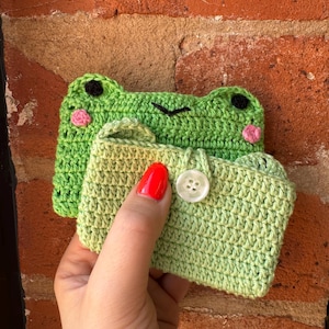 May include: Handmade crochet frog-shaped coin purse in shades of green, with black eyes and pink cheeks. The purse has a small handle and a button closure. A second frog-shaped item is visible behind the purse.