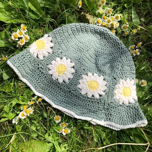 Può includere: Un cappello a secchiello verde all'uncinetto con fiori di margherita bianchi. Il cappello ha un bordo bianco all'uncinetto attorno al bordo.