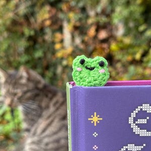 May include: A handmade green crochet frog bookmark with black eyes and pink cheeks sits atop a purple book. The frog is made of yarn and has a smiling face. The book has gold star and floral designs.
