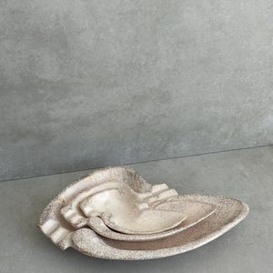 May include: A set of three ceramic dishes in a nesting arrangement. The dishes are off-white with a speckled, textured finish. The largest dish has a unique, organic shape, while the smaller ones fit inside.