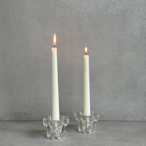 May include: Two lit, ivory-colored taper candles in clear glass candle holders. The candle holders have a decorative, floral-like design. The background is a solid, light gray color.