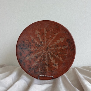 Puede incluir: Un plato decorativo redondo, de color marrón rojizo, con un diseño geométrico. El plato presenta un patrón central de rayos de sol con elementos triangulares y lineales. El plato se exhibe sobre un soporte de metal plateado sobre un fondo neutro.
