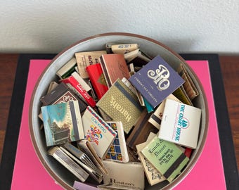May include: A collection of vintage matchbooks in a round, gray bowl with a brown rim. The matchbooks are of various colors and designs, with some featuring text like "Harrah's" and "The Chart House". The bowl sits on a pink surface.