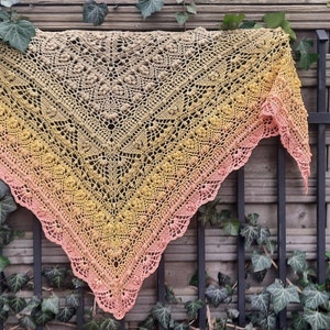 Può includere: Uno scialle all'uncinetto con un gradiente di colori rosa, giallo e beige. Lo scialle è drappeggiato su una recinzione di legno con edera che cresce su di essa.