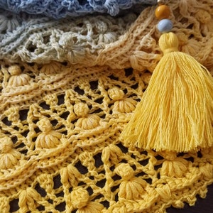 Può includere: Una coperta all'uncinetto gialla con un fiocco di nappe. La coperta è realizzata con un delicato motivo a punto e presenta un gradiente di colori dal blu chiaro al giallo.