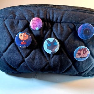 May include: A black quilted cosmetic bag with six colorful pinback buttons attached. The buttons feature various designs, including a cartoon character, a cat, and floral and abstract patterns. One button has the text "Fairest of Them All Boutique."