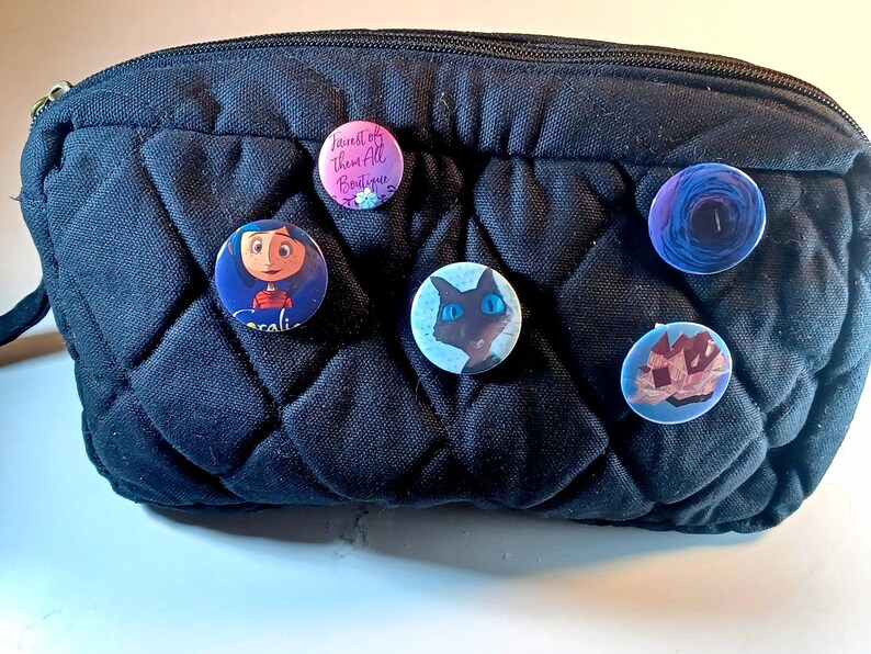 May include: A black quilted cosmetic bag with six colorful pinback buttons attached. The buttons feature various designs, including a cartoon character, a cat, and floral and abstract patterns. One button has the text "Fairest of Them All Boutique."