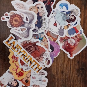 May include: A collection of colorful stickers featuring characters and imagery from the movie "Labyrinth." The stickers include images of David Bowie, owls, and other fantasy elements. The word "LABYRINTH" is printed in gold letters.