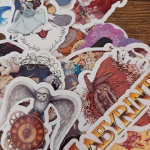May include: A collection of fantasy-themed stickers featuring characters and imagery from the movie Labyrinth. The stickers include images of an owl with clockwork, a couple dancing, and the word "LABYRINTH" in gold lettering. The stickers are on a wooden surface.