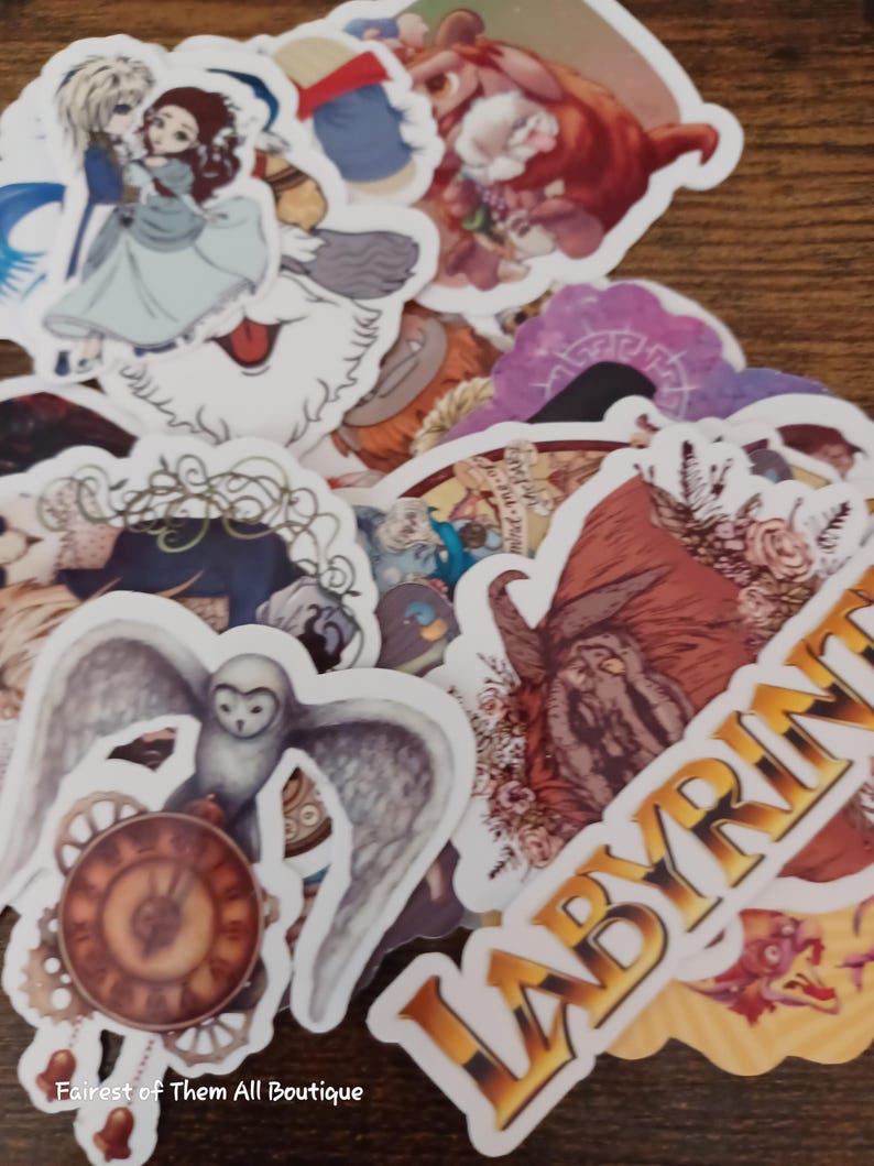 May include: A collection of fantasy-themed stickers featuring characters and imagery from the movie Labyrinth. The stickers include images of an owl with clockwork, a couple dancing, and the word "LABYRINTH" in gold lettering. The stickers are on a wooden surface.