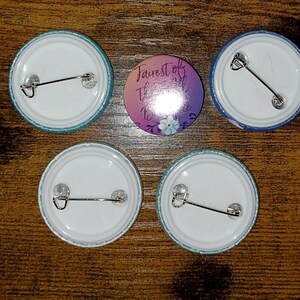 May include: Five round pinback buttons. Four buttons have a white center and a teal or blue border. One button has the text "Fairest of them all" with a floral design in purple and blue.