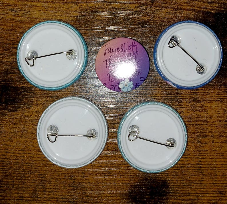 May include: Five round pinback buttons. Four buttons have a white center and a teal or blue border. One button has the text "Fairest of them all" with a floral design in purple and blue.