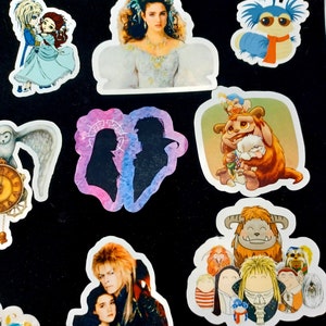 May include: A collection of colorful stickers featuring characters from the movie Labyrinth. The stickers include images of Jareth the Goblin King, Sarah, Hoggle, and other iconic characters. The stickers are on a black background.