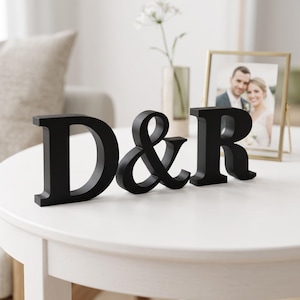 May include: A freestanding black decorative sign featuring the uppercase letters D and R separated by an ampersand symbol. The letters are crafted with a bold serif font and have a smooth matte finish, designed to stand upright on a flat surface.