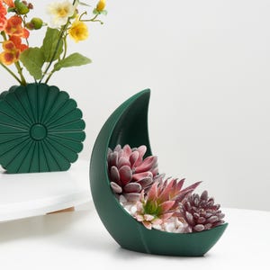 Puede incluir: Una jardinera verde oscuro en forma de media luna llena de suculentas artificiales y guijarros blancos. Un jarrón verde a juego con un arreglo floral está en el fondo. Las flores son naranjas, amarillas y blancas.