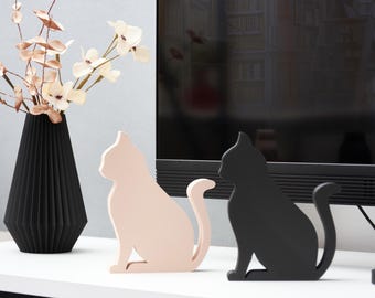 May include: Two wooden cat figurines, one black and one light pink, sitting on a white surface in front of a black television.