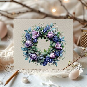 K&ouml;nnte beinhalten: Eine Karte mit einem Aquarellkranz aus lila und blauen Blumen und gr&uuml;nen Bl&auml;ttern. Der Kranz befindet sich auf einem beigefarbenen Hintergrund. Die Karte ist von Ostereiern und einer Feder umgeben.