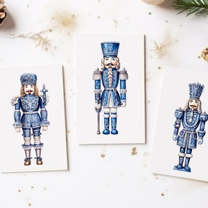 May include: Three blue and white nutcracker figurines with gold accents. The nutcrackers are standing on white cards.