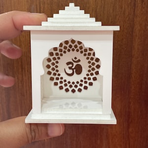 Puede incluir: Un pequeño templo de madera blanco con un corte decorativo del símbolo Om. El templo tiene un techo escalonado y una pequeña plataforma en el interior.