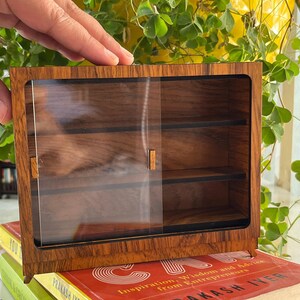 Op de afbeelding: Een klein houten kastje met glazen deuren en planken. De kast is gemaakt van donker hout met een natuurlijke houtnerf. De glazen deuren zijn helder en laten de inhoud van de planken zien.