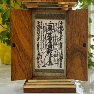 Può includere: Un espositore in legno con parte superiore e base color oro, con una pergamena incorniciata con calligrafia nera. La custodia ha due ante a cerniera e una base rettangolare. Il legno ha una venatura visibile.