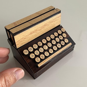 Puede incluir: Una máquina de escribir de madera en miniatura con un acabado marrón y veta de madera. Las teclas están etiquetadas con las letras del alfabeto y tienen un aspecto vintage.