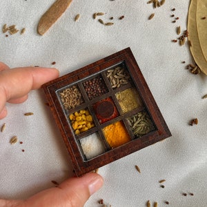 Puede incluir: Una caja de especias de madera con nueve compartimentos, cada uno lleno de una especia diferente. Las especias incluyen comino, cilantro, semillas de mostaza, cúrcuma, chile en polvo, semillas de hinojo y sal. La caja está hecha de madera oscura y tiene una tapa de vidrio transparente.