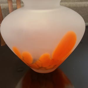 May include: A frosted glass vase with a rounded body and a flared rim. The vase features orange, petal-like designs near the base. The vase is likely used for decorative purposes, such as holding flowers or other items.